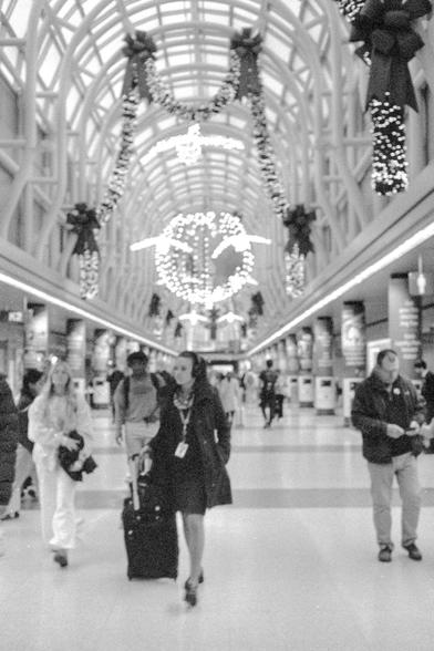 Black and white photo of people walking down a great hallway in an airport with Christmas decorations and lights under a glass arch atrium.