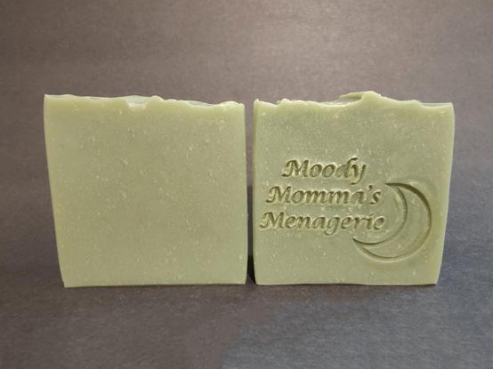 Two earthy green colored soaps sitting side by side. The bar on the right is stamped with my branding "Moody Momma's Menagerie" and a crescent moon.