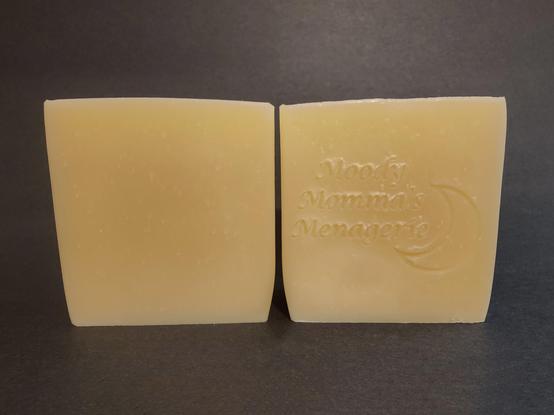 Two cream colored soaps sitting side by side. The bar on the right is stamped with my branding "Moody Momma's Menagerie" and a crescent moon.