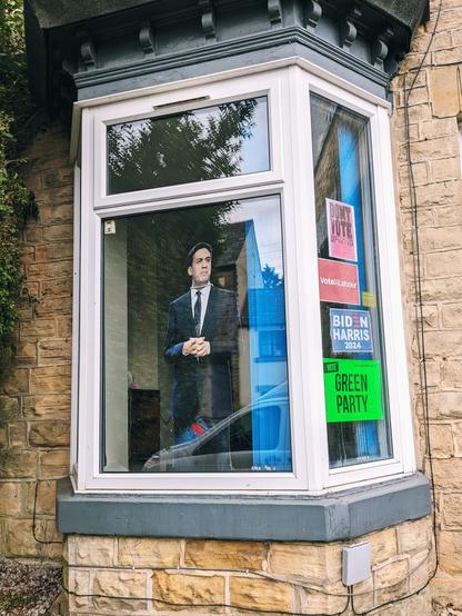 Picture of the front bay window of a Victorian era terraced house. The right side has several political posters reading (from top to bottom):
Don't Vote 
Vote Labour 
Biden Harris 2024
Green Party 

In the main part of the window, a cardboard cutout of Ed Miliband can be seen, wearing a (presumably fake) pearl necklace.