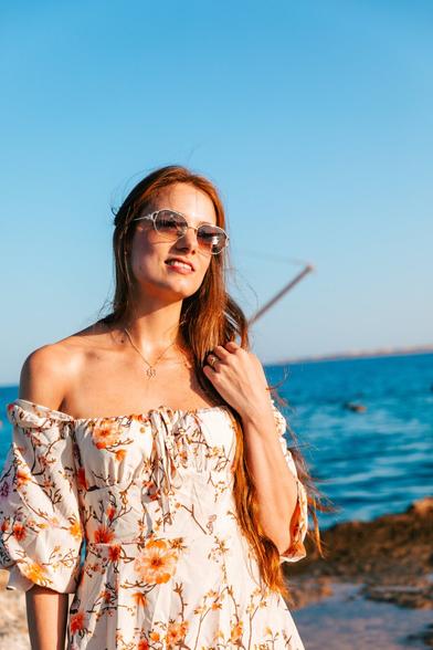 A young woman wearing sunglasses and a floral dress, standing by the sea with a relaxed pose.
