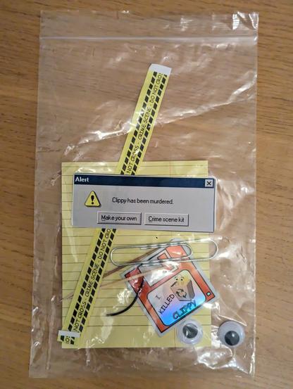 A plastic bag containing parts to make a Clippy murder crime scene, with a large paperclip, googly eyes, eyebrows, yellow card and "CRIME SCENE DO NOT CROSS" tape, along with an "I killed Clippy" sticker.