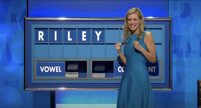 TV show screenshot of Rachel Riley, the polymathematically-skilled cohost of Countdown, in front of the "letters" board she manages during the TV game show "Countdown".  She has just drawn the 5th out of 9 letters in the "word game" and randomly spelled her first name, eliciting a celebratory pair of fists and grin, taking the opportunity to look into the studio camera.  (At this moment the studio audience gives a large cheer).