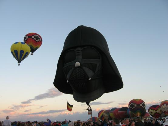 A picture of the Dark Vader hot air balloon launching at the Albuquerque International Balloon Fiesta.  Numerous other hot balloons can be seen airing up and getting ready to launch.  Several other hot air balloons can be seen in the sky.