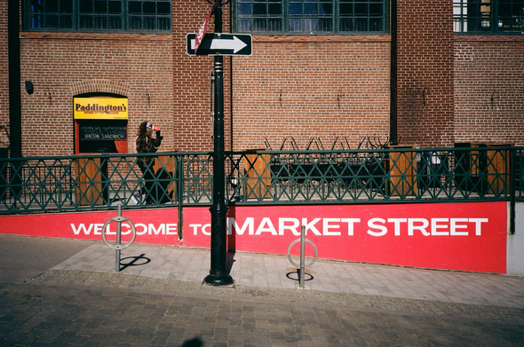 A concrete retaining wall is emblazoned, in whit block letters on a bright red background, “Welcome to Market Street”. On the walkway above a pedestrian, drinking coffee as they walk, has just passed a window advertising “Paddington’s, Home of the Oink”. They takeout coffee cup is also bright red with white script.