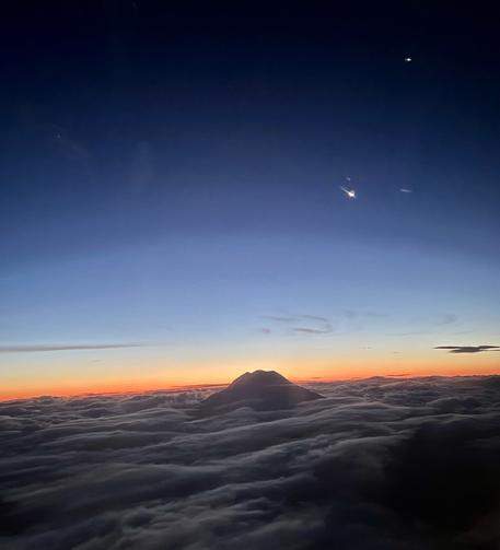 A photo taken from inside an airplane window of Mt Rainier emerging from a bed of clouds, our moon, and in the same line, Jupiter. A seam of orange sunlight is just emerging from the horizon and the sky is awash with saturated dark blues.