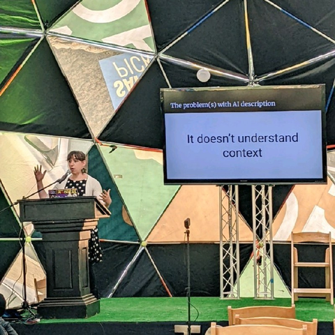 Me behind a lectern on a stage motioning with my hands, a slide on a large screen behind me reads "the problem(s) with AI description: it doesn't understand context"
