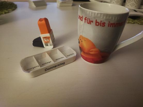 Eine große Tasse mit Kaffee und dem Schriftzug "Dich lieb ich am liebsten und für bis immer" von der Firma Tatzino, ein weiße Pillendose für Donnerstag und ein Asthma Spray auf einen weißen Wohnzimmertisch.