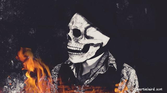 Monochrome still frame from Dimittit In Artes with a man in a skull mask and beret sitting in a dark wet shed surrounded by flames.