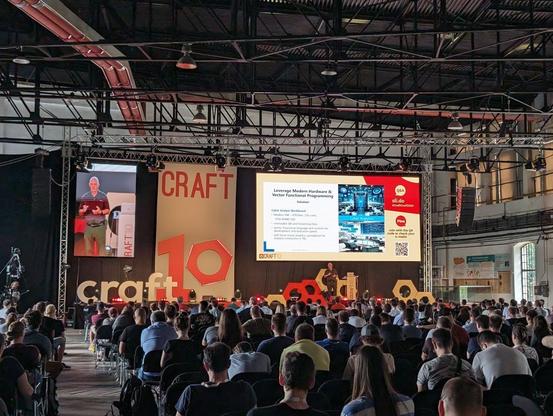 Blick von hinten auf die Bühne des Konferenz-Saals bei der Craft Conference in Budapest. Der Saal ist voll mit Besucherinnen und Besuchern. Auf der Bühne steht ein Speaker und hält einen Vortrag.