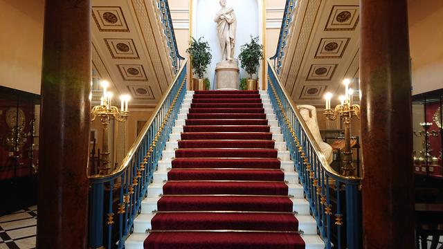 A fancy staircase inside the Town Hall leading up to a half-landing with a marble statue flanked by two potted shrubs