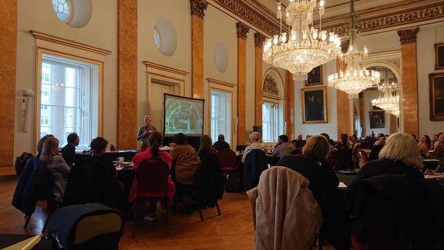 A man presenting to a room of people sat around circular tables.  He's stood in front of a projector screen showing an aerial view of a road network for kids.

The room has a row of sash windows along one wall, with circular windows above each of those.  It has inset columns at regular intervals, topped with ornate Corinthian reliefs and is lit by elaborate glass chandeliers.