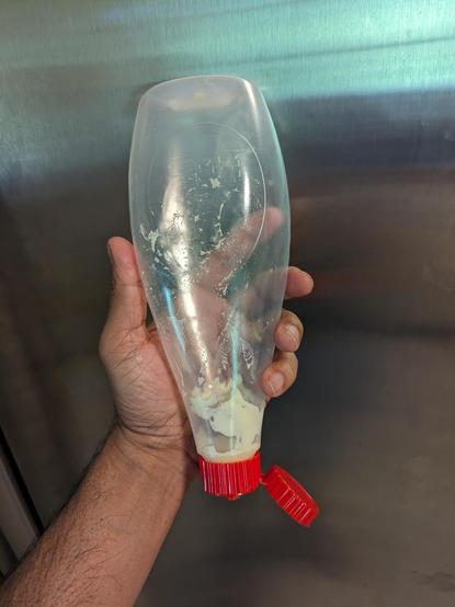 A plastic bottle of Kewpie mayonnaise held upside down.  The bottle is almost clear plastic except for a bit of mayonnaise just near the red bottle cap