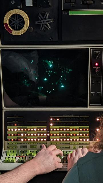 Spacewar! is being played on the PDP-12's scope. A starfield is shown. Two spaceships and a hail of bullets fly around a central star -- all drawn on a green phosphorescent screen with phosphor streaks. The PDP-12 green, black, and white console is lit up like a Christmas tree. Hands are on the right switches, controlling the switches.