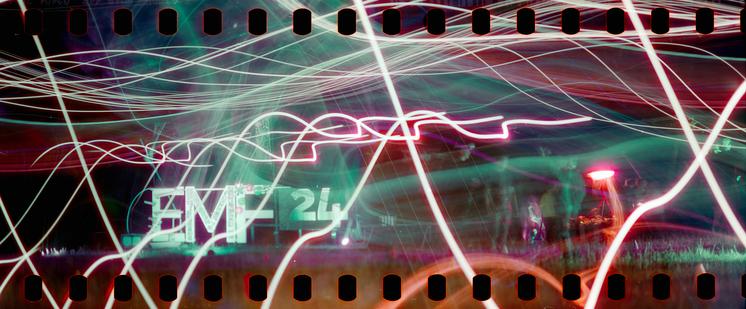 Panoramic film photograph with sprocket holes visible above and below. Subject is a large lit up sign reading "EMF24". There are many light streaks in abstract pattern running across the image.