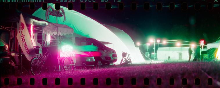 Panoramic film photo with sprocket holes visible above and below. Picture is of a nightly scene with a neon sign "FRESH ROBOTS" on a pushcart illuminating tents around it.