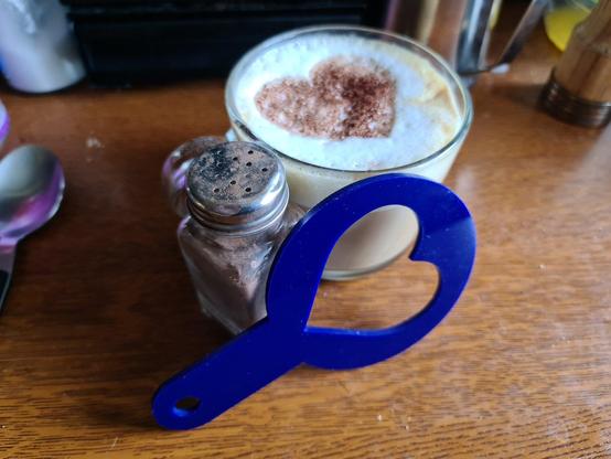 A glass coffee cup with cocoa powder heart in the top of the foamed milk. In front of the cup is the plastic stencil used to make the heart shape, and a dusting pot with cocoa power in.