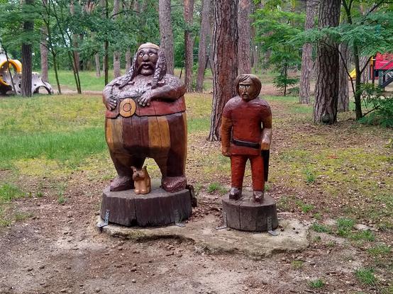 Wooden figures on two tree stumps.

The left figure is a male with a huge belly. On it, a wide red belt with gold-colored buckle, and below striped trousers, with interleaved wide brown and yellow stripes. His face is round with a friendly look, with a flat cap (or a helmet?) on top, wide mustache and hair in two braids falling onto his chest. His hands are resting on his belly. There is a small, bright brown dog at his feet, probably a terrier.

The right figure is a smaller, thin male in a red garment. He's wearing a sword belt. His face is slender, with wide unibrow, narrow lush mustache and short lush hair.

In the background, you can see trees in a park. To the sides, fragments of children's playground structures.