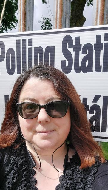 A pale white woman with auburn hair wearing a black top and sun glasses, stands in front of a sign saying Polling Station.