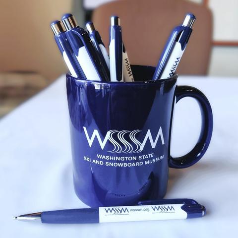 A blue and white WSSSM branded ballpoint pen rests quietly before a blue WSSSM mug full of other ballpoint pens