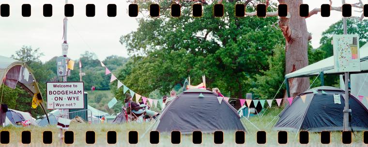 Panoramic film photograph with sprocket holes visible above and below. Photo of two tents on a field, next to them is a traffic sign which reads "Welcome to BODGEHAM ON WYE - Wye not?". A large tree and more tents on the background.