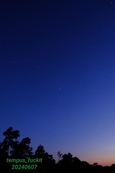 Fotografie (eine Sekunde belichtet) des Himmels kurz nach dem Sonnenuntergang. In der rechten unteren Ecke ist noch der orangene Schein des Restlichts der Sonne zu sehen. Je weiter links oben im Bild, desto mehr dominieren dunklerwerdende Blautöne. Zwischendrin erkennt man Sterne. Am unteren Rand und in der rechtsunteren Ecke sind schemenhaft Baumkronen zu erkennen. Rechts unten befindet sich auch mein Wasserzeichen im üblichen giftgrün.