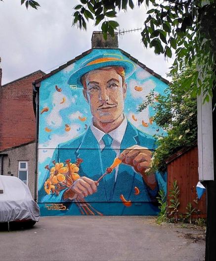 Streetartwall. The mural of a man picking flowers is sprayed on the outside wall of a single-storey beige house. The background of the mural is a turquoise blue sky with white clouds over which yellow petals swirl. The red-haired man with the narrow moustache is wearing a turquoise suit, tie, hat and white shirt. He is holding a bouquet of yellow flowers and plucking petals with both hands.
Note: The mural is reminiscent of figures from paintings by Van Gogh, Gauguin or Paul Signac. A modern version of various elements of the painter's friends. The yellow flowers, the title "A la Folie", the red-haired man and the blue sky. But perhaps just a coincidence.