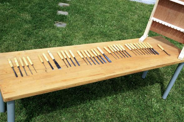 langer Holztisch auf dem 35 Schnitzmesser aufgereiht sind, dazu ein Eigenbau-Holzständer, in dem die Schnitzmesser in zwei Reihen zur Aufbewahrung eingehängt werden können.