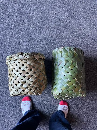 Two baskets woven from Harakeke leaves. They are on their sides, my feet are shown below them for scale.