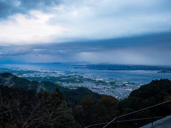 A panoramic view of a coastal city with buildings and greenery, surrounded by a large body of water and mountainous terrain under an overcast sky.