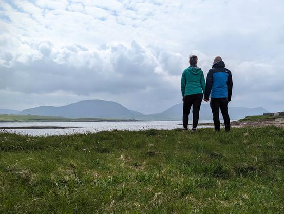 A photo taken from just above ground level of a couple facing away, holding hands, looking across a narrow passage of sea (the Hoy Sound) towards an island (Hoy) with a couple of hills on it. There is grass in the foreground; another, flat, island (Graemsay) to the left; and a rockier section with an old lookout and shed to the right of the couple. The couple are a woman on the left wearing a bright green raincoat and a man on the right wearing a bright blue raincoat with navy shoulders and sleeves.