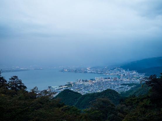 A coastal city with surrounding mountains and forested areas under a cloudy sky.
