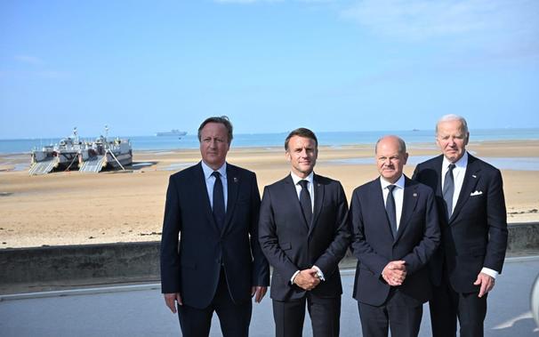 A Normandy beach (Sword, Juno, Gold, Omaha or Utah). Two modern Landing Craft Vehicle Personnel (LCVP) are beached above the waterline. An amphibious warfare ship lurks on the horizon. On the seafront stand four men in suits (combat kit for politicians). On our right is President of the United States Joe Biden, to his right is Chancellor Scholz of Germany, French President Macron stands to his right. On Macron's right is, er, the Untied Kingdom's Foreign Secretary, Baron Cameron of Greensill. Sorry, Lord Cameron of Huggahoodie. Strangely yet appropriately Dave's face is hidden in shadow.