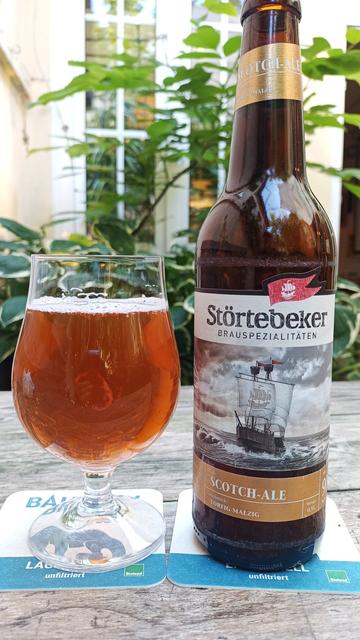 Auf einem Holztisch steht ein bauchiges Bierglas mit rötlich braunem Bier gefüllt. Daneben steht die Bierflasche mit Aufschrift:
Störtebeker Scotch Ale