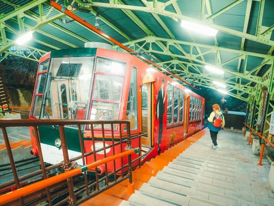 A red and green funicular train at a station, with a person wearing a red backpack walking down the stairs beside it. The station has a green metal roof structure and bright lighting.