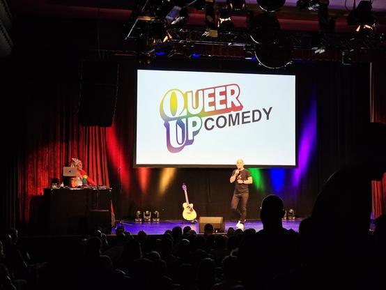 Markus Barth und DJ Lana Delicious auf der Bühne vom Gloria in Köln bei QueerUp Comedy. Die Name der Veranstaltung ist groß auf einem Banner sichtbar in Regenbogen Farben. Musikinstrumente stehen im Hintergrund. Bunte Lichter strahlen in Regenbogen Farben die Rückwand an.