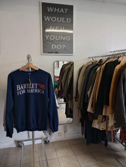 a Barlett '98 for America sweatshirt hangs on the front of a rack of clothes, a large sign reads WHAT WOULD NEIL YOUNG DO on the wall.  a rack of leaather jackets is on the right