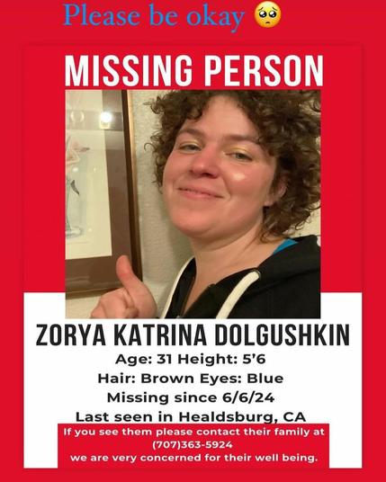 A missing person poster with a red background features a photograph of a woman with short, curly brown hair, giving a thumbs up and smiling slightly. The top of the poster reads "Please be okay" with a sad emoji. Below the photo, the text reads:

MISSING PERSON

ZORYA KATRINA DOLGUSHKIN

    Age: 31
    Height: 5'6"
    Hair: Brown
    Eyes: Blue
    Missing since: 6/6/24
    Last seen in Healdsburg, CA

If you see them, please contact their family at (707) 363-5924. We are very concerned for their well-being.