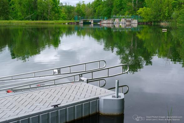 A grey floating dock with shiny silver rails extends from the lower left corner of the frame into a lake reflecting trees and sky. In the distance, three orange buoys are suspended in front of a concrete dam and sluice gate.