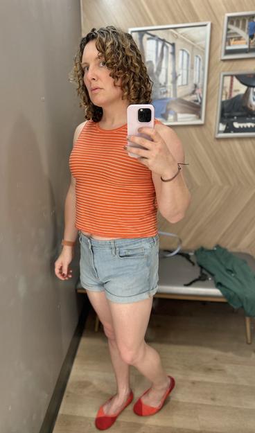 Me, a white trans woman with a vaguely irritated expression, in a changing room. I’m wearing an orange and white-striped shirt and jean shorts with red/coral shoes.