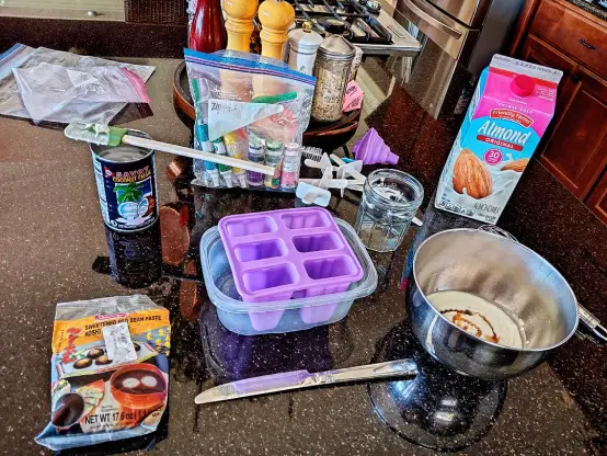 various ingredients to make Asian style popscicles. Popscicle mold in purple, red bean paste, a silver bowl with white and tan mixture, almond milk carton, a baggie of flavored extracts, can of coconut creme with a spatula on top.