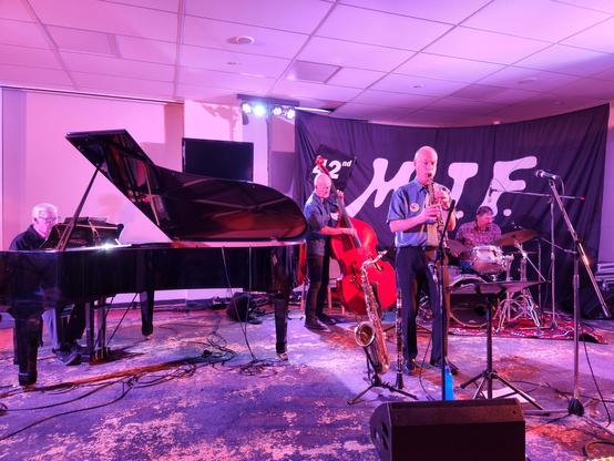 Four blokes... Piano on left, then bass, clarinet, and Ken on drums, all on a stage with pinkish lighting.