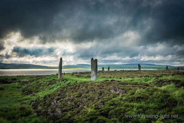 Photo of a partial circle of standing stones on a grassy platform surrounded by a ditch. The sky is dark and ominous but the sun is breaking through in the background and shining on a loch.