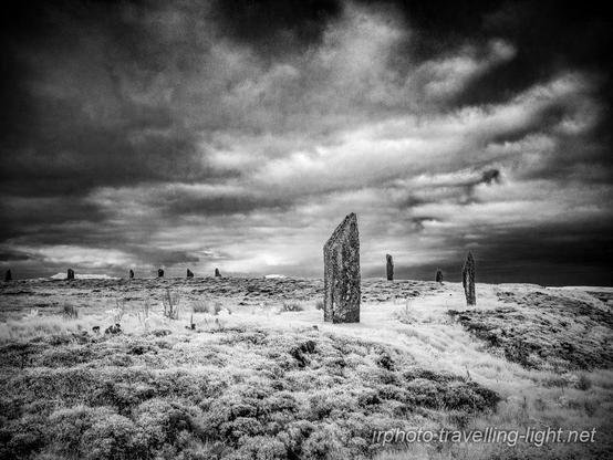Black and white infrered photo of a partial circle of standing stones on a grassy platform surrounded by a ditch, with a dramatic sky.