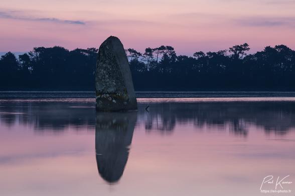 Photo, en couleur présentant un menhir cerné par la marée haute, au lever du jour. L'ambiance colorée est plutôt  pourpre, le soleil n'étant pas encore levé. Le menhir se reflète dans l'eau, très calme, et son sommet dépassent légèrement, la cime des pins de l'autre rive de la rivière, l'appareil photo, ayant été placé relativement près du sol pour avoir cette perspective et mettre en valeur le menhir.