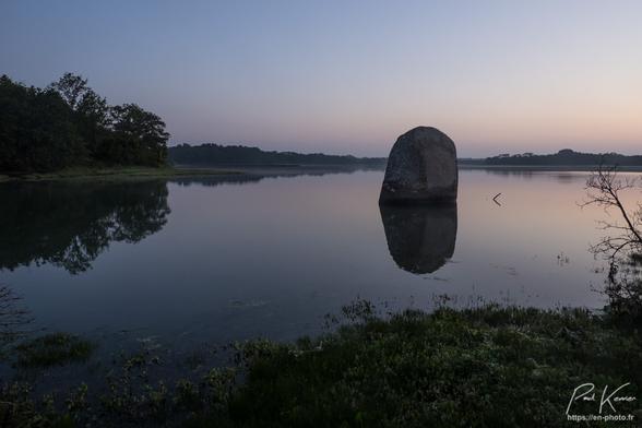 Photo, en couleur présentant un menhir cerné par la marée haute, au lever du jour, sous un angle différent avec donc un profil plus large du menhir. L'ambiance colorée est plutôt  bleue, le soleil n'étant pas encore levé. Le menhir se reflète dans l'eau, très calme. Au bas de la photo, on voit la végétation de la rive, d'où a été prise la photo, alors qu'en haut de la photo derrière le menhir, envoie la végétation de la rive opposée avec des arbres à gauche, et une rive nettement plus éloignée derrière le menhir et sur la droite de la photo.