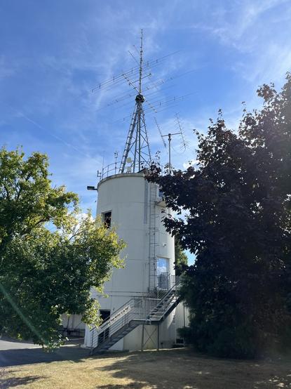 Zu sehen ist das Clubheim des OV N47 Gütersloh. Ein Betonsilo mit großer Antennenanlage auf dem Dach. Im Vordergrund Bäume und im Hintergrund blauer Himmel mit strahlendem Sonnenschein.