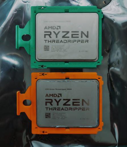 Two AMD Threadripper CPU's placed on a silvery antistatic surface. Top one is a 2950x with a green bracket, bottom one is a 1900x with an orange bracket.