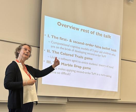 Rineke Verbrugge introducing her class on modal logic and social cognition. She stands in front of a slide, containing the text:

Overview rest of the talk:

• 1. The first- & second-order false belief task
— Computational cognitive models of 5 year old children who are on the brink of developing second-order ToM

• Il. The Colored Trails game
— Using a software agent to entice students' theory of mind

• III The Marble Drop game
— What makes applying second-order ToM in a turn-taking game so difficult?