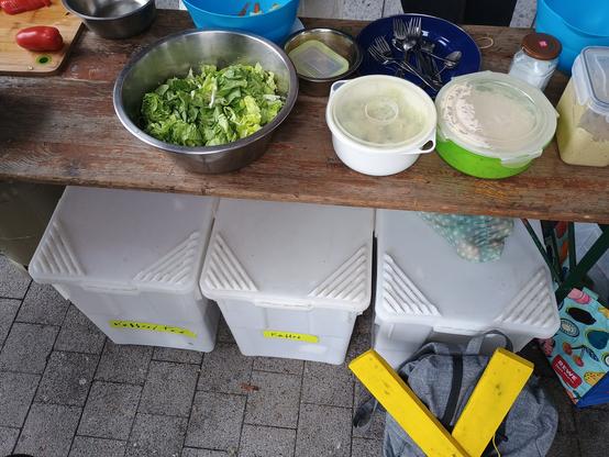 Foto von Biertisch mit Salatschüsseln und weissen Boxen am Boden. Auch ein gelbes X an Rucksack ist zu sehen.